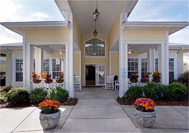 Entrance of a senior living facility named Summit Place with a covered walkway, white rocking chairs on the porch, hanging flower pots, and potted plants with orange and purple flowers on either side of the walkway.