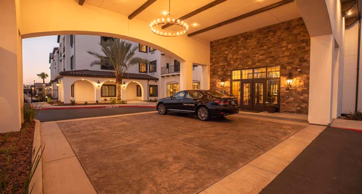 Entrance area of The Park At Laguna Springs facility at dusk, showing a covered driveway with a black car parked under a large archway. The building features stone and stucco walls, warm lighting, and palm trees wrapped with string lights.