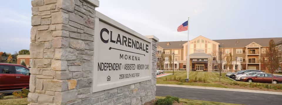 Exterior view of Clarendale of Mokena senior living facility with a stone sign in the foreground displaying the facility name and services offered. The building is in the background with an American flag on a flagpole and several parked cars visible.