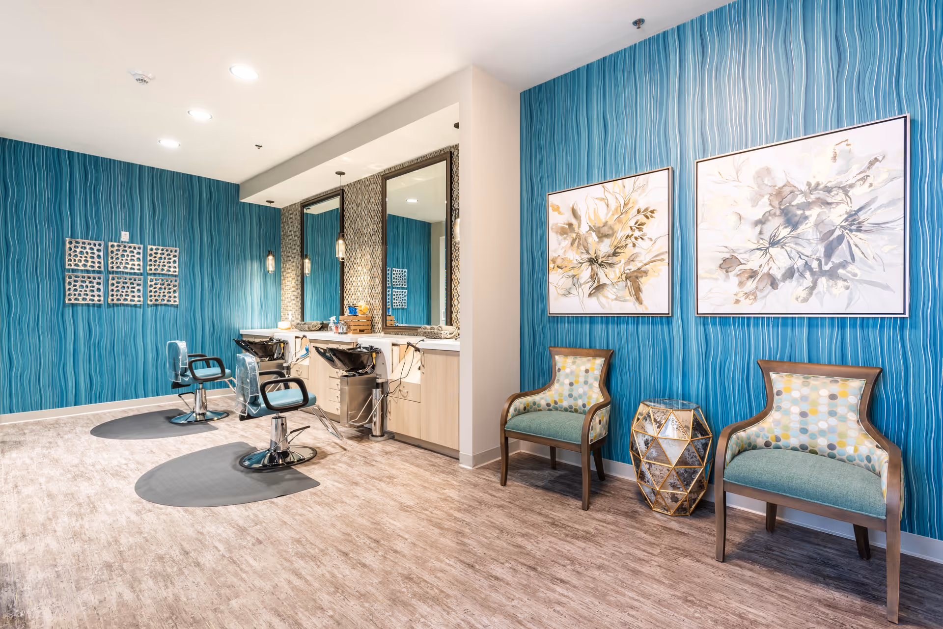 Bright salon area with turquoise patterned walls, two styling chairs and wash sinks, two upholstered guest chairs and framed floral artwork.