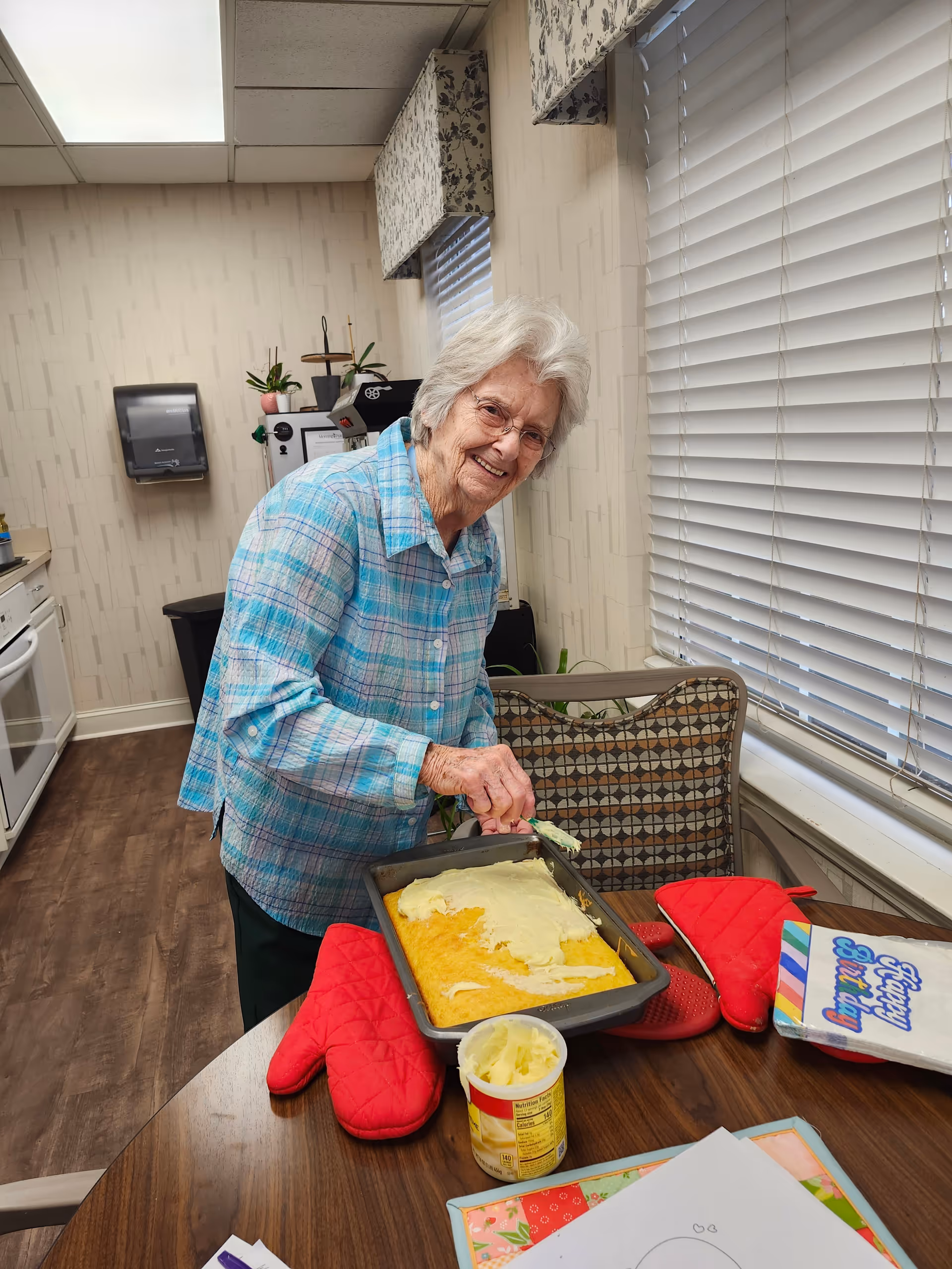 An elderly woman with white hair and glasses is smiling while spreading frosting on a rectangular cake in a kitchen. She is wearing a blue plaid shirt. The kitchen has wooden floors, white cabinets, and a window with blinds. Red oven mitts and a container of frosting are on the table along with some papers and a colorful birthday card.