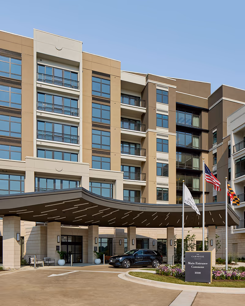 Exterior view of The Carnegie at Washingtonian Center senior living facility showing the main entrance with a covered drop-off area, a black car parked in front, three flagpoles with flags, and a sign indicating the main entrance and commons. The building is multi-story with large windows and a modern design under a clear blue sky.