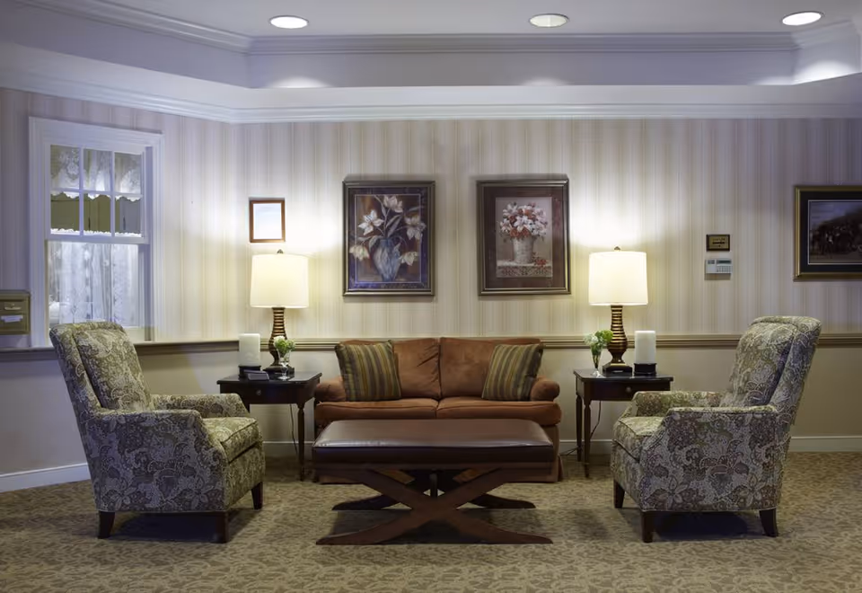 A cozy living room area with a brown sofa flanked by two patterned armchairs. There are two side tables with lamps and small plants on either side of the sofa. The walls have striped wallpaper and are decorated with framed floral artwork. A window with lace curtains is visible on the left side.