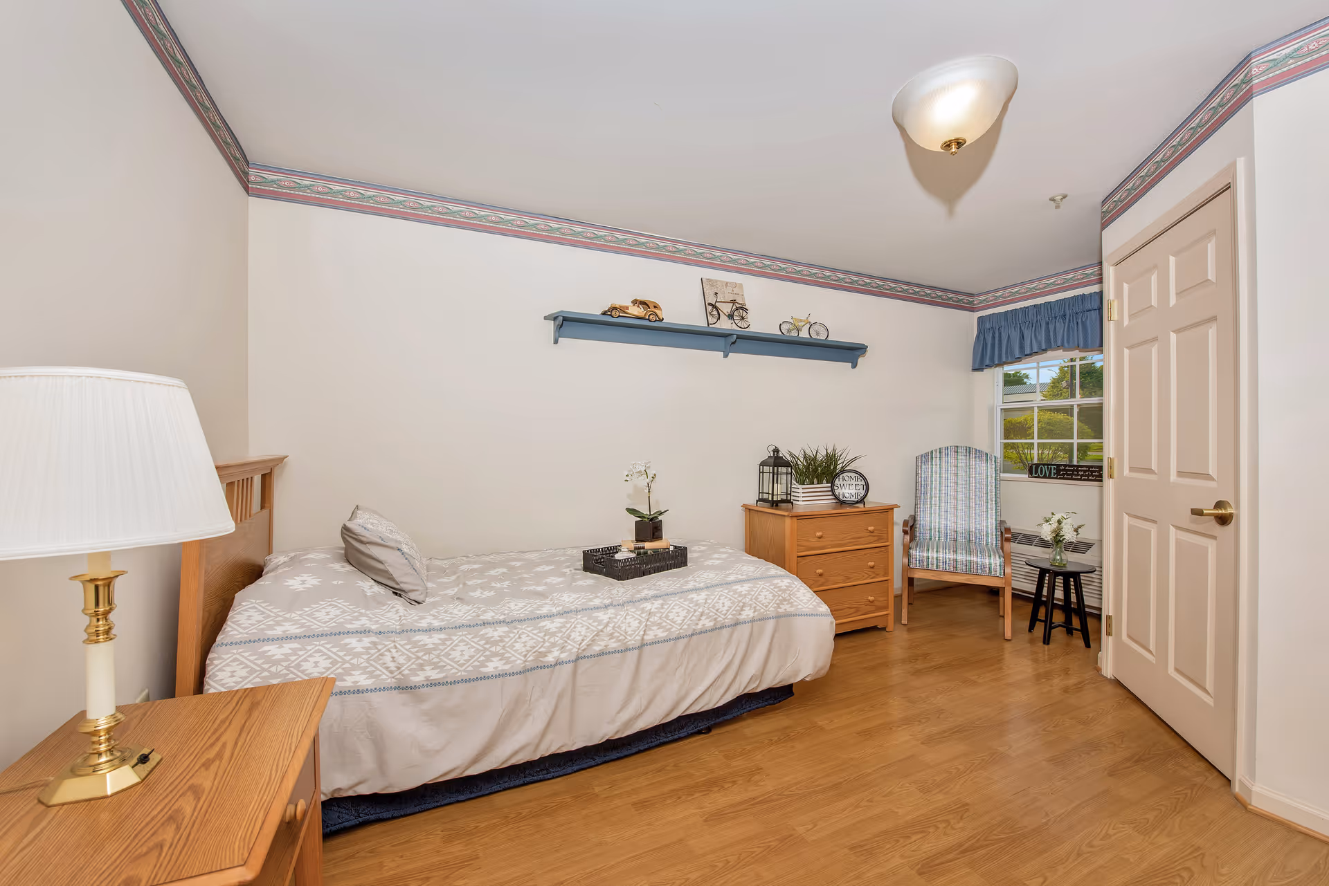 A cozy bedroom with a single bed covered in a patterned beige and white bedspread. There is a wooden nightstand with a lamp on the left side of the bed. On the right side of the room, there is a wooden dresser with decorative items including a plant and a lantern. Above the dresser, a blue wall shelf holds small decorative objects. A cushioned chair with a striped pattern is placed near a window with blue valance curtains. The room has light-colored walls with a decorative border near the ceiling and wood flooring.