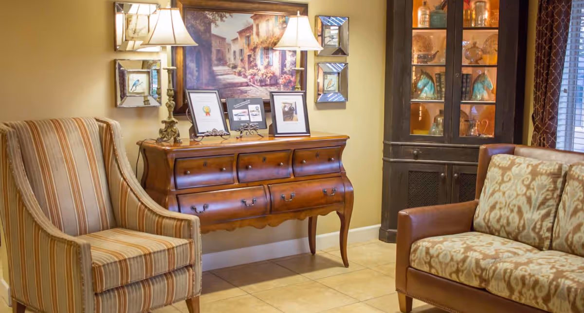 A cozy sitting area with a striped armchair on the left and a patterned sofa on the right. Between them is a wooden console table with decorative lamps, framed pictures, and a large painting of a street scene hanging above it. To the right is a dark wooden cabinet with glass doors displaying decorative items.