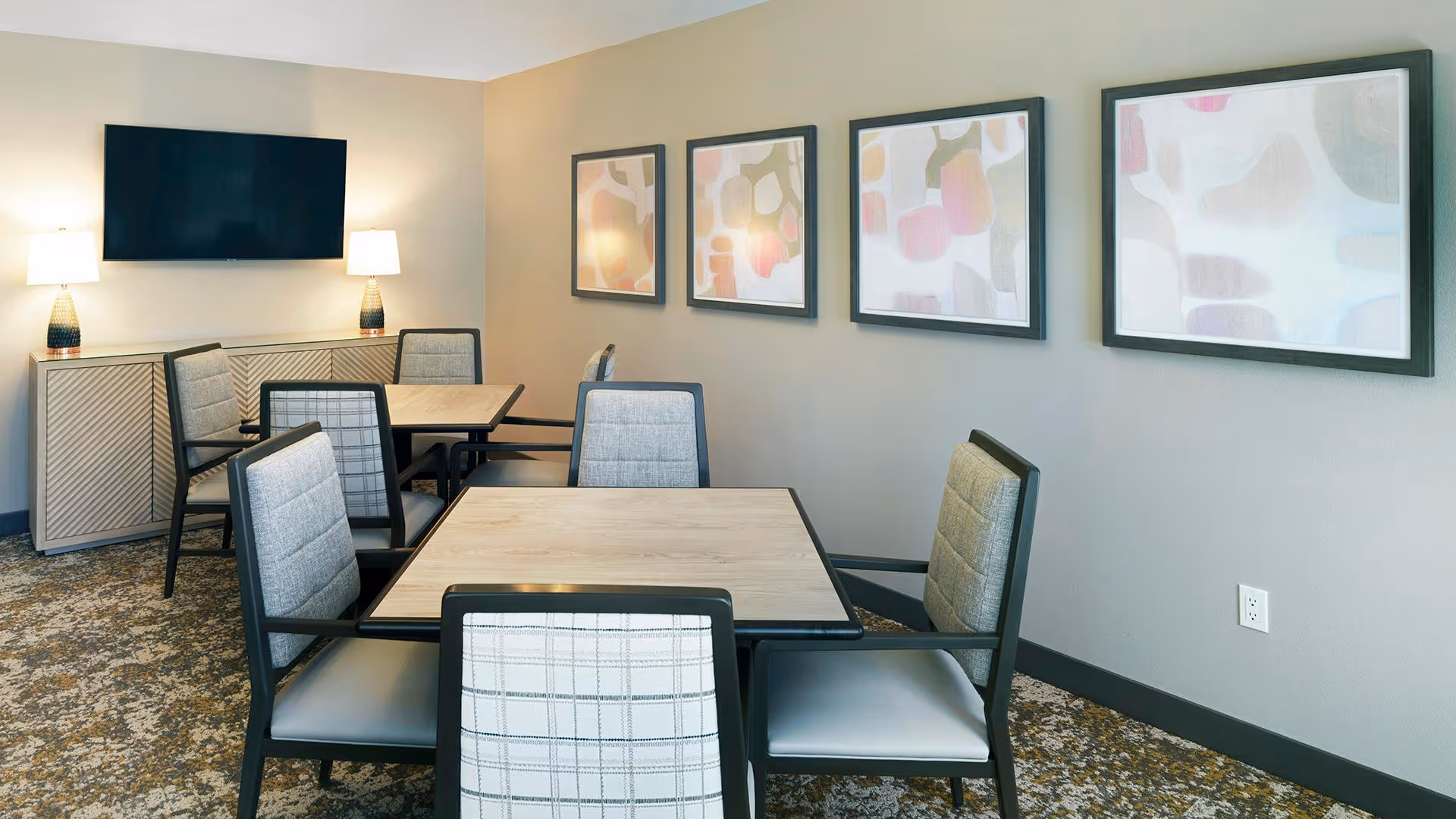 A common area with two square tables surrounded by cushioned chairs with armrests. The room has a patterned carpet, light gray walls adorned with four abstract art pieces, a flat-screen TV mounted on the wall above a sideboard, and two table lamps providing warm lighting.