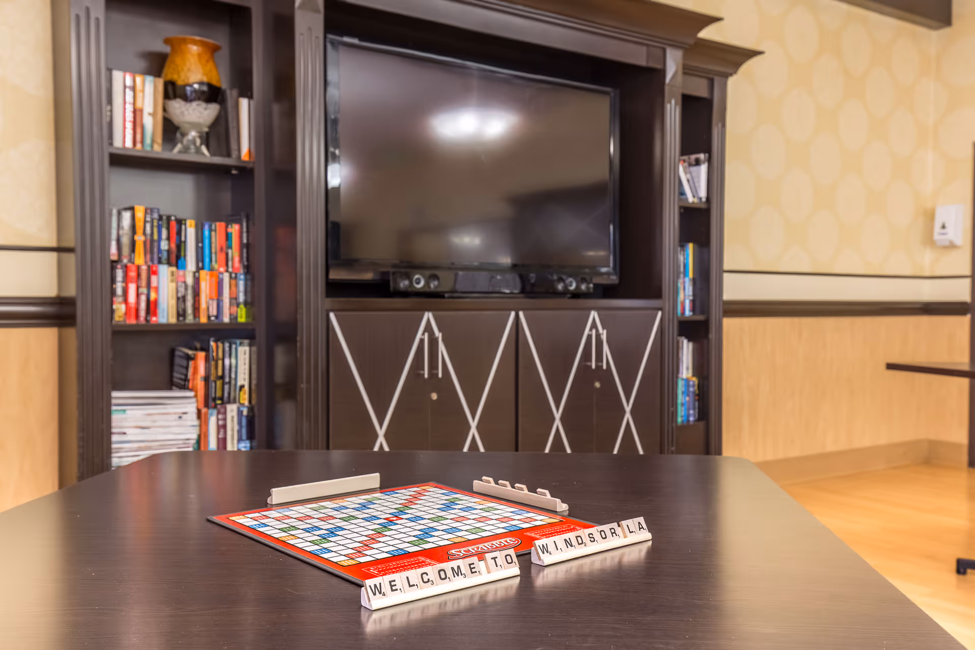 A cozy common area with a dark wooden table in the foreground featuring a Scrabble board game with tiles spelling out 'WELCOME TO WINDSOR LA'. In the background, there is a large flat-screen TV mounted on a dark wooden entertainment center with shelves filled with books and decorative items. The room has warm-toned walls and wooden flooring.