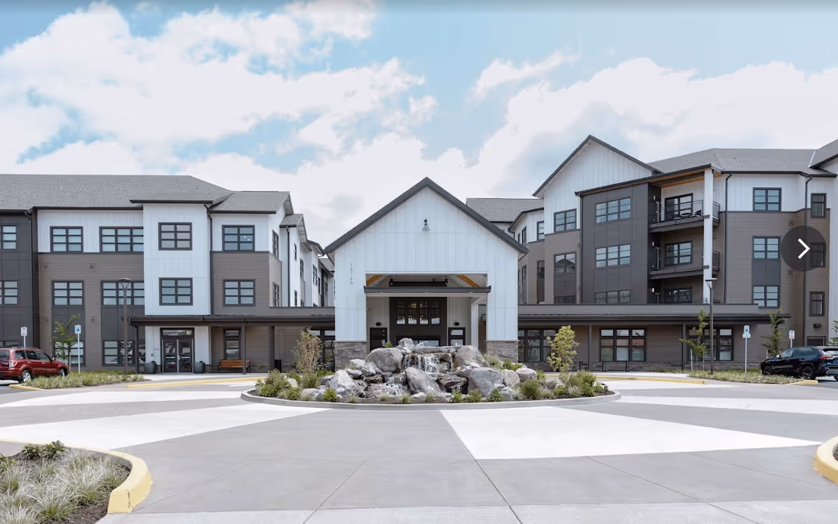 Front exterior view of The Springs at Happy Valley facility showing a modern multi-story building with gray and white siding, large windows, a central entrance with a peaked roof, and a landscaped roundabout with rocks and a small waterfall feature.
