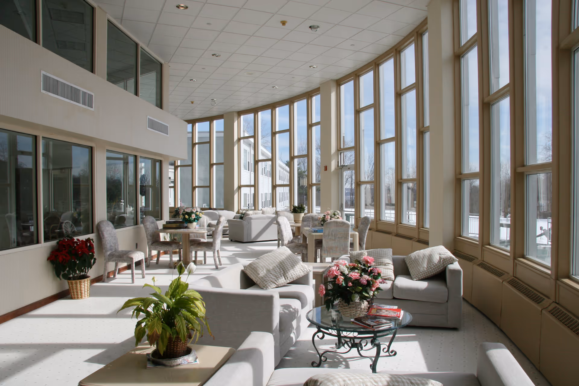A bright and spacious common area with large floor-to-ceiling windows letting in natural light. The room is furnished with comfortable armchairs, sofas, small tables with floral arrangements, and dining tables with chairs. There are plants placed around the room, and the view outside shows a snowy landscape.