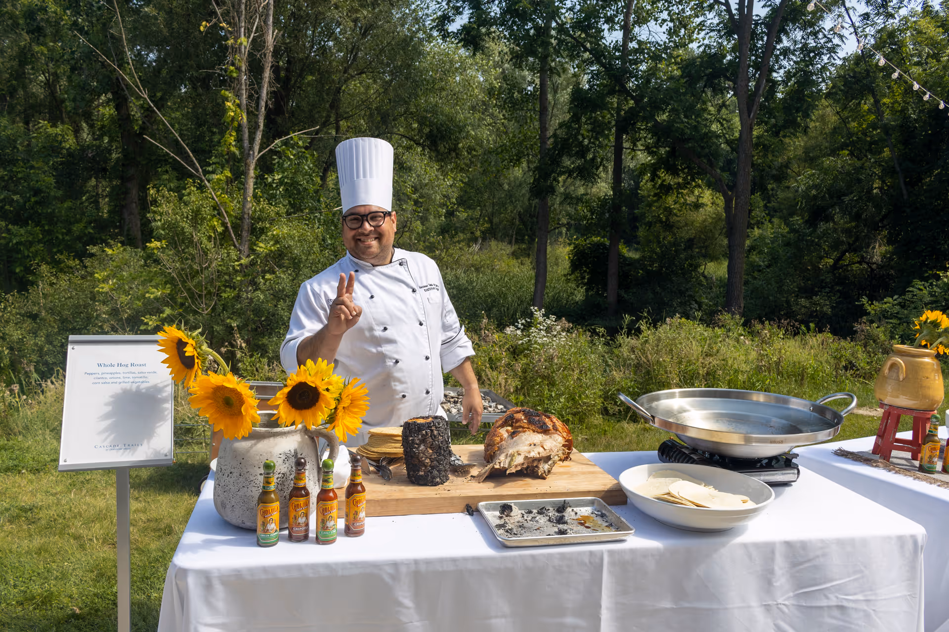 A chef wearing a white uniform and tall white hat stands behind a table outdoors with a cutting board holding a roasted whole hog. The chef is smiling and making a peace sign with one hand. The table is covered with a white tablecloth and has sunflowers in a vase, several bottles of hot sauce, a bowl with tortillas, a large pan, and a sign describing the whole hog roast. The background shows green trees and grass under a sunny sky.