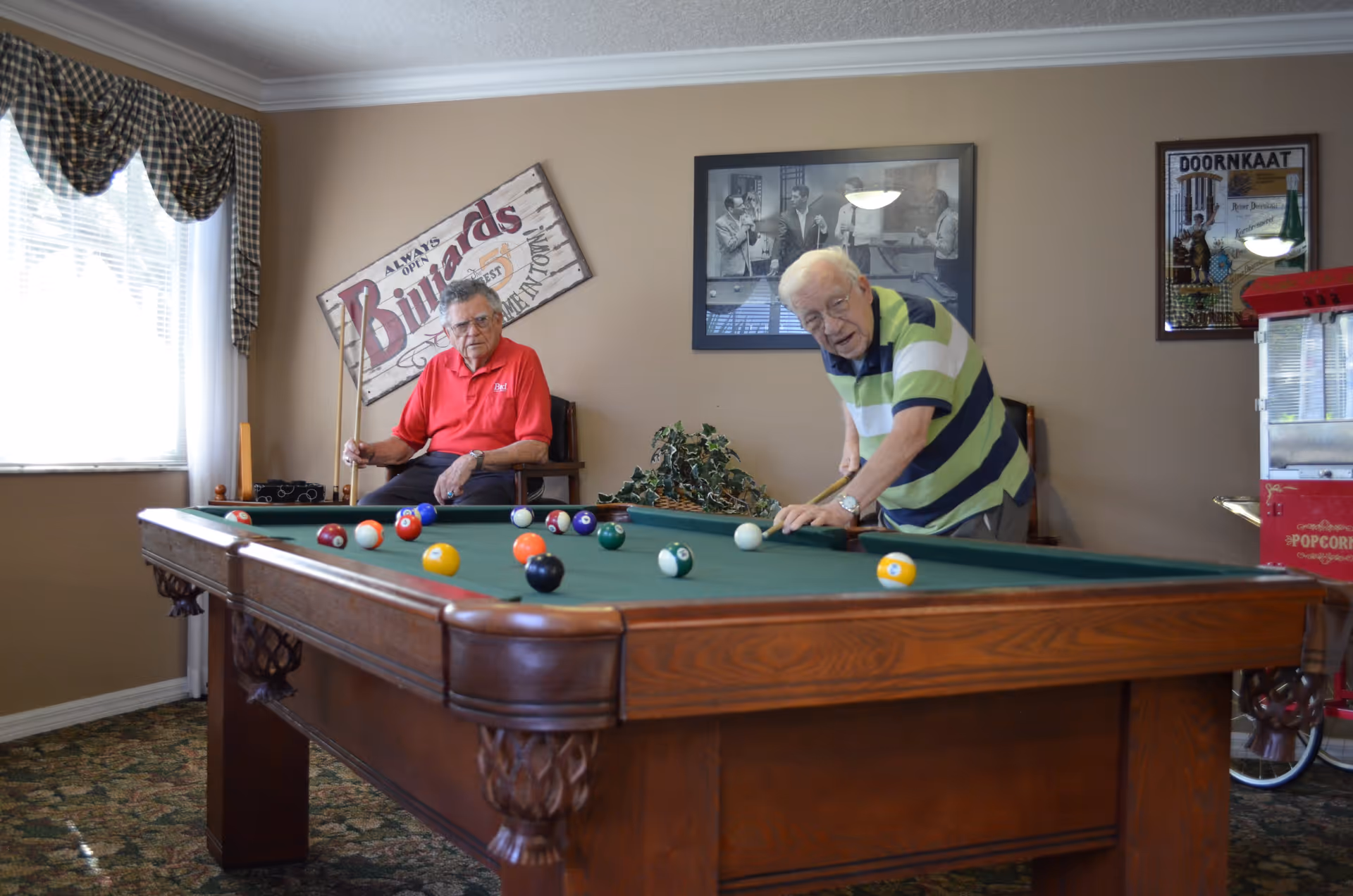 Two elderly men playing billiards in a recreational room with a pool table. One man in a red shirt is sitting on a chair holding a cue stick, while the other man in a green and navy striped shirt is leaning over the table preparing to take a shot. The room has beige walls with framed pictures and a sign that reads 'Billiards'. There is a window with checkered curtains and a popcorn machine visible on the right side.