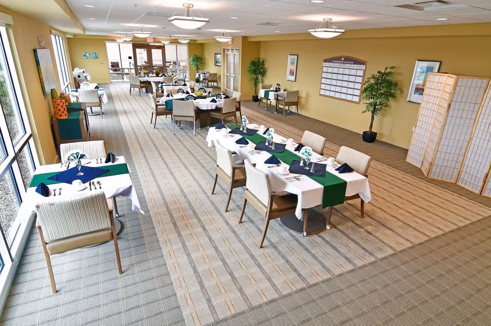 A spacious dining room in a senior living facility with multiple tables set with white tablecloths, green and navy blue runners, napkins, cups, and silverware. The room has large windows on one side, beige walls, carpeted floors, potted plants, framed artwork, and a large calendar on the wall. There is a folding screen in one corner and ceiling lights providing illumination.