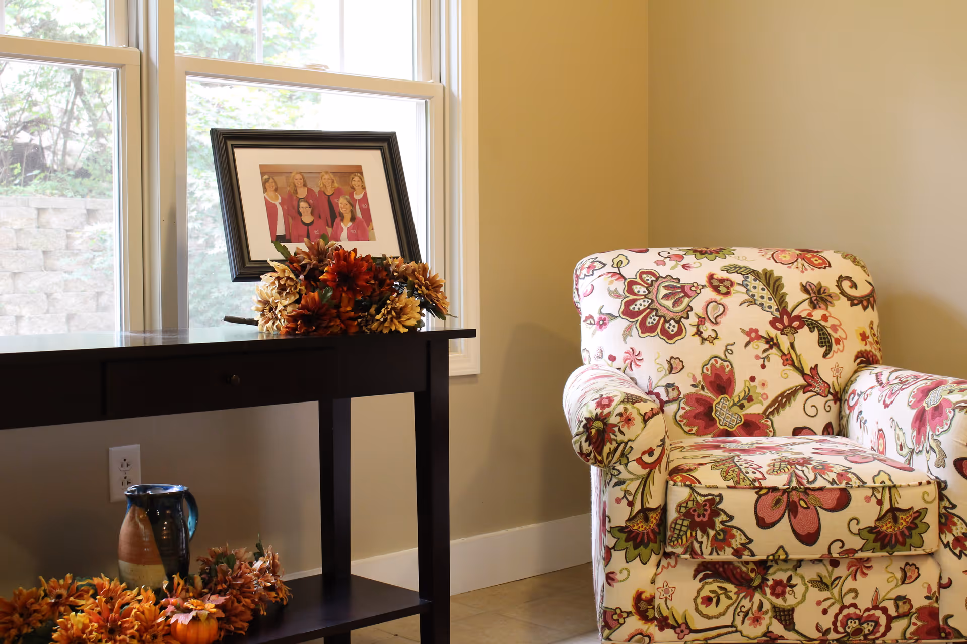 A cozy corner of a room featuring a floral-patterned armchair next to a dark wooden table. The table holds a framed photo of six women and a bouquet of autumn-colored flowers. Below the table is a ceramic pitcher and more autumn-themed decorations. A window with white trim lets in natural light, and a stone wall is visible outside.