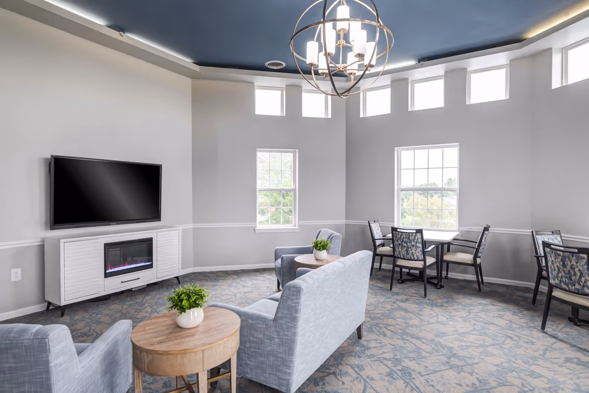 A bright and spacious common area with light gray walls and a blue ceiling accent. The room features a wall-mounted flat-screen TV above a white cabinet with an electric fireplace. There are two light gray upholstered armchairs and a matching loveseat arranged around a round wooden coffee table with a small green plant. In the background, there are two windows letting in natural light and two small tables with patterned chairs. A modern spherical chandelier hangs from the ceiling.