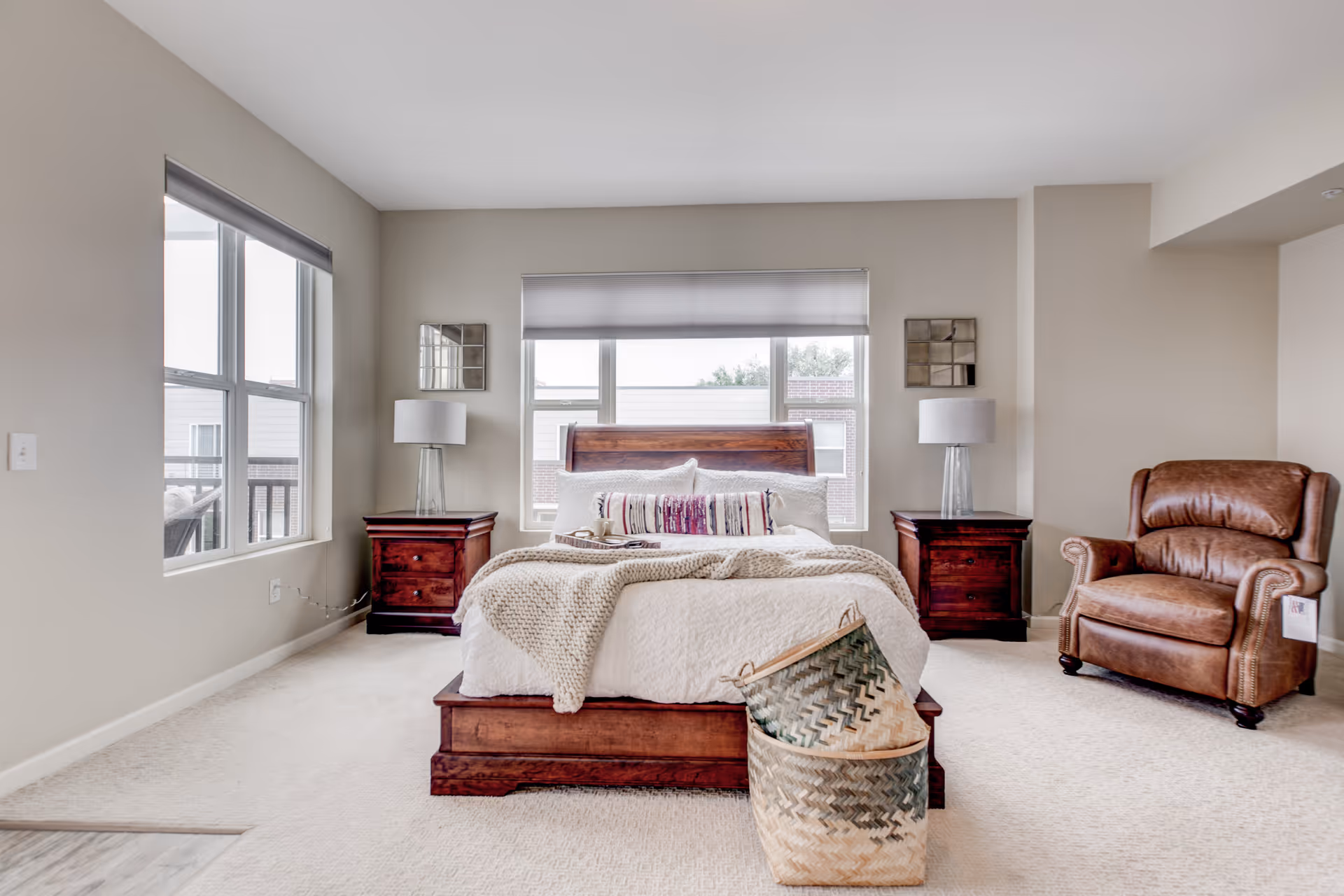 A bright and spacious bedroom with a large wooden bed centered against a wall with three windows. The bed is made with white bedding and decorative pillows. There are two matching wooden nightstands on either side of the bed, each with a modern lamp. To the right, there is a brown leather armchair. In front of the bed, there are two woven baskets stacked together on a light carpeted floor. The walls are painted a soft beige color.