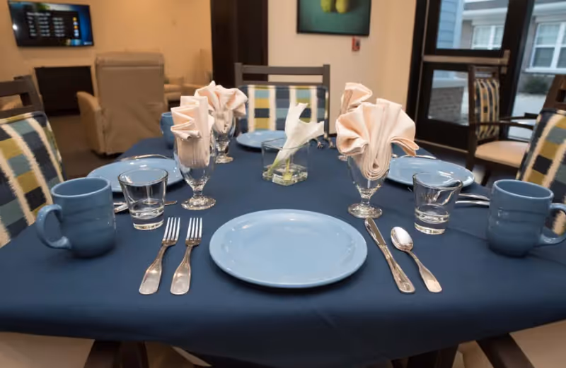 A dining table in a communal dining room set with blue plates, mugs, glassware, folded napkins and a small flower centerpiece.