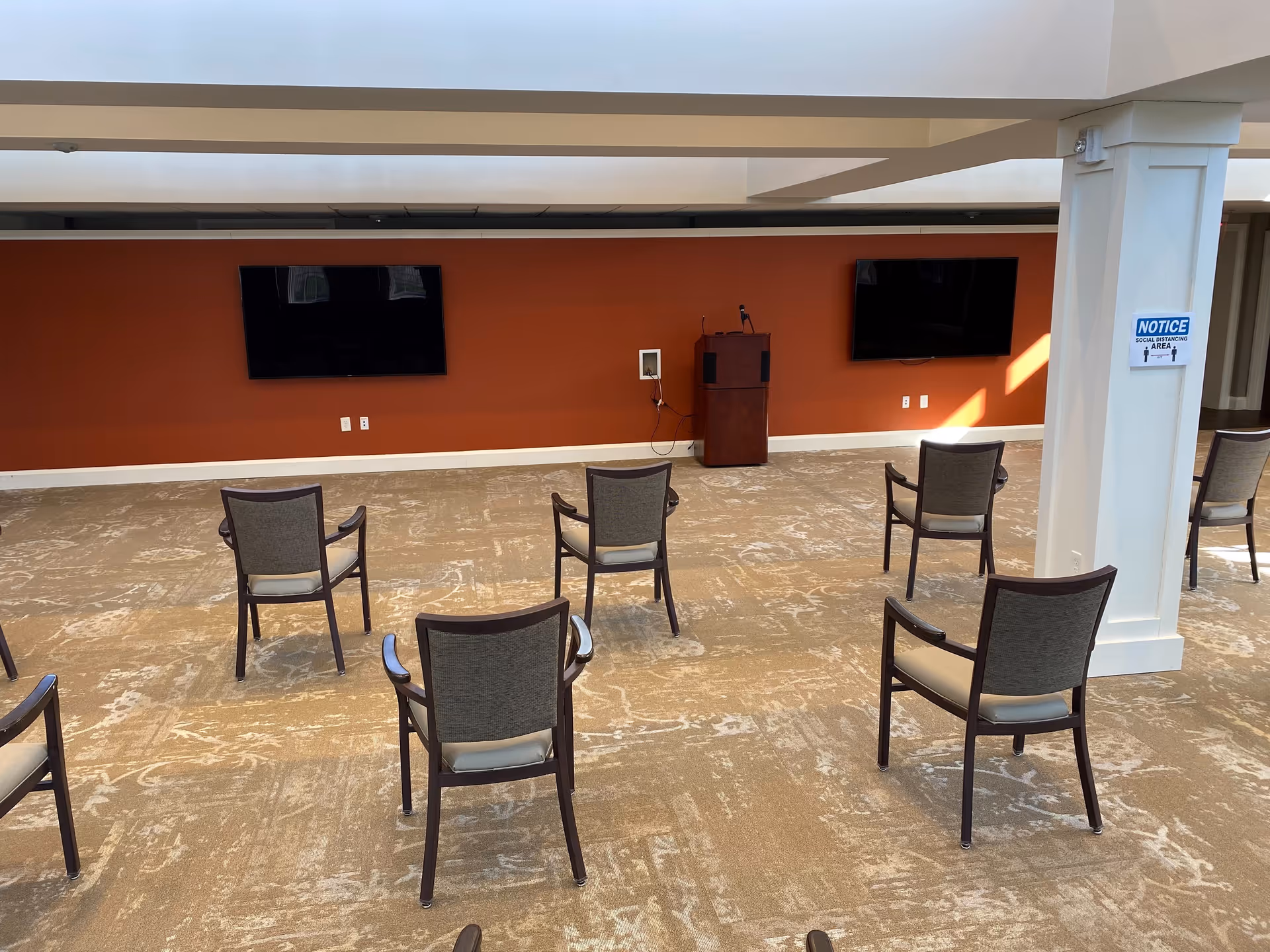 A spacious room with a beige patterned carpet and an orange accent wall featuring two large flat-screen TVs mounted side by side. Several chairs are arranged in rows facing the TVs, spaced apart for social distancing. A wooden podium with a microphone is positioned between the TVs. A white pillar on the right side has a blue and white notice sign indicating a social distancing area.