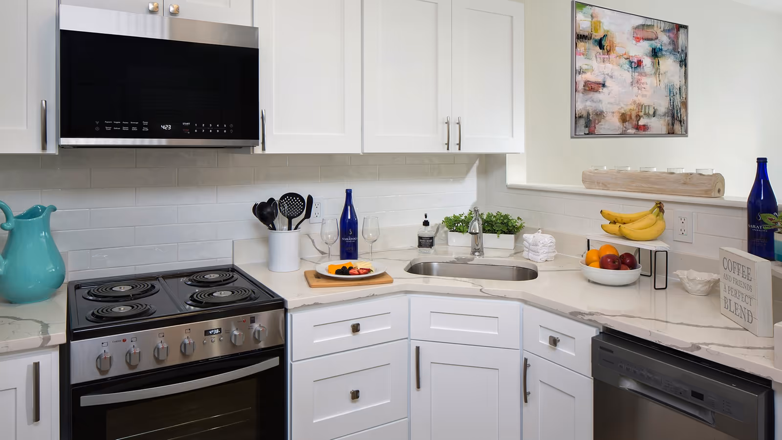 A modern kitchen with white cabinets and marble countertops. There is a stainless steel stove with four burners and an oven, a microwave above it, a sink with a faucet, and a dishwasher. On the counter, there are kitchen utensils in a white holder, a cutting board with sliced fruit, a blue bottle, a bowl of bananas, a bowl of mixed fruit, a small plant, a soap dispenser, a stack of white towels, and a decorative sign that reads 'COFFEE AND FRIENDS A PERFECT BLEND'. A colorful abstract painting hangs on the wall.