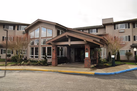 Exterior view of Brookdale Wilsonville senior living facility showing a three-story building with large windows and a covered entrance supported by brick columns. The surrounding area includes landscaped bushes and trees, a paved driveway, and a sidewalk with a blue curb.