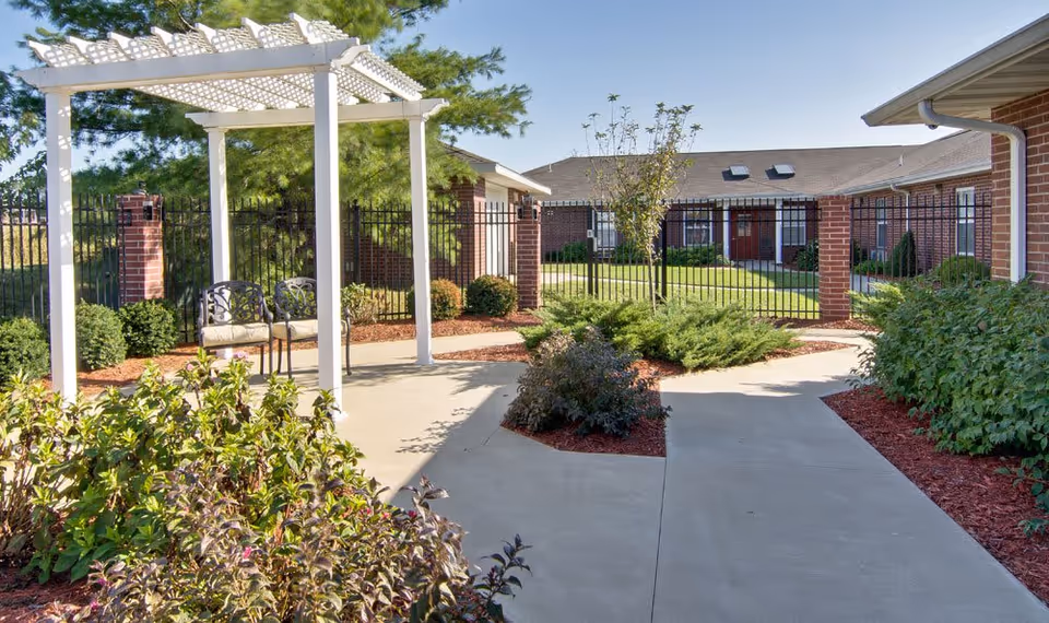 Outdoor courtyard area with a white pergola, two metal chairs with cushions, surrounded by green bushes and plants. The courtyard is paved with concrete walkways and enclosed by a black metal fence. Brick buildings with windows and doors are visible in the background under a clear blue sky.