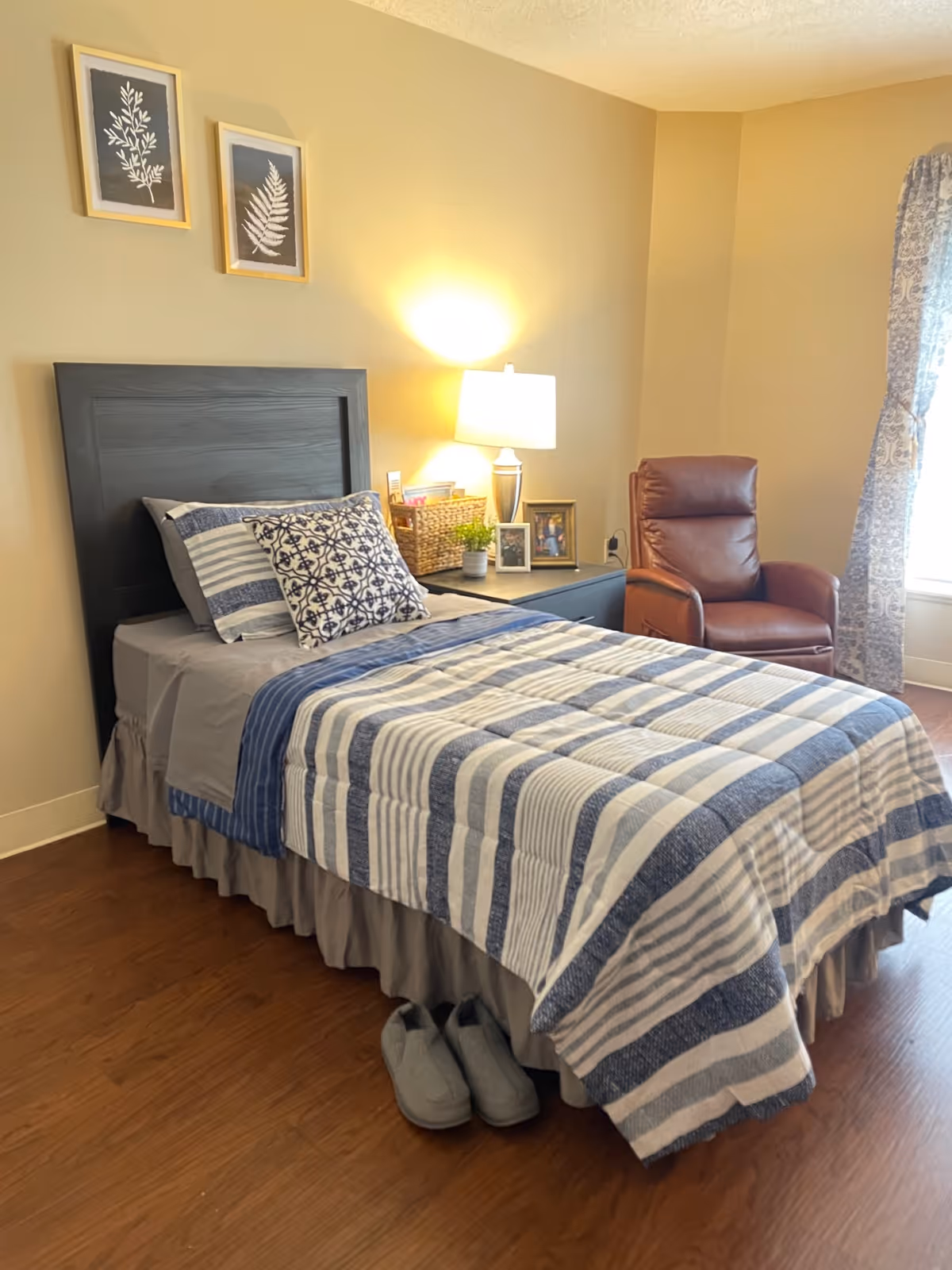 A cozy bedroom with a single bed covered in a blue and white striped quilt and matching pillows. Next to the bed is a nightstand with a lamp, framed photos, a small plant, and a basket. A brown leather armchair is positioned near a window with patterned curtains. The floor is wooden, and a pair of gray slippers are placed at the foot of the bed. Two framed botanical prints hang on the beige wall above the bed.