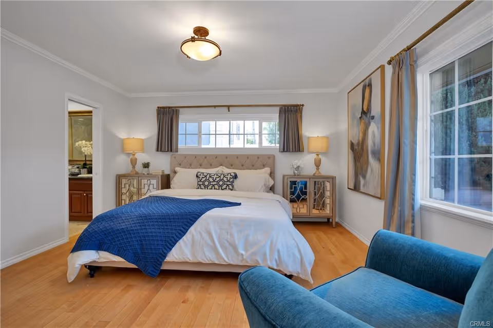 A bright bedroom with a large bed covered in white bedding and a blue throw blanket. There are two matching bedside tables with lamps on each side of the bed. A window with curtains is above the bed, and another window with curtains is on the right wall. A blue armchair is positioned near the window on the right side. A doorway on the left leads to a bathroom with a sink and mirror visible. The room has light wood flooring and white walls with a large abstract painting on the right wall.