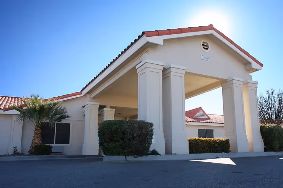 Exterior view of a senior living facility building with a covered entrance supported by large white columns, red-tiled roof, and surrounding bushes and palm tree under a clear blue sky.