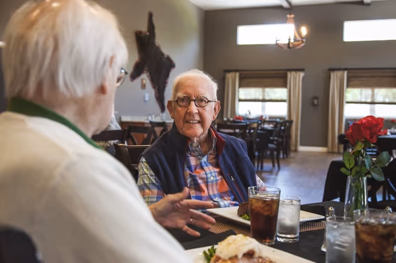 Two elderly men sitting at a dining table in a well-lit room, engaged in conversation. One man is wearing glasses and a plaid shirt with a navy vest, while the other is seen from behind. The table has plates of food, glasses of iced tea and water, and a vase with red roses. The background shows more dining tables and windows with curtains.