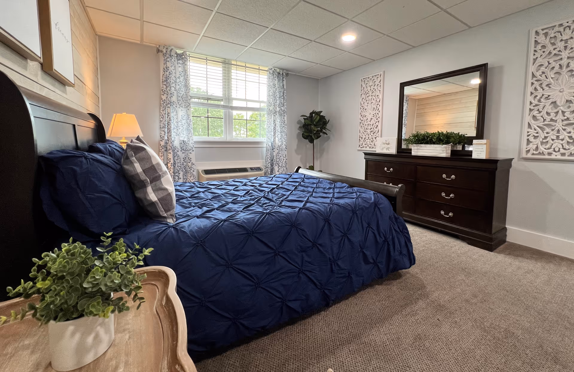A bedroom with a dark wooden bed frame and a navy blue comforter, a checkered pillow, a wooden dresser with a large mirror, decorative wall art, a potted plant, and a window with patterned curtains letting in natural light.