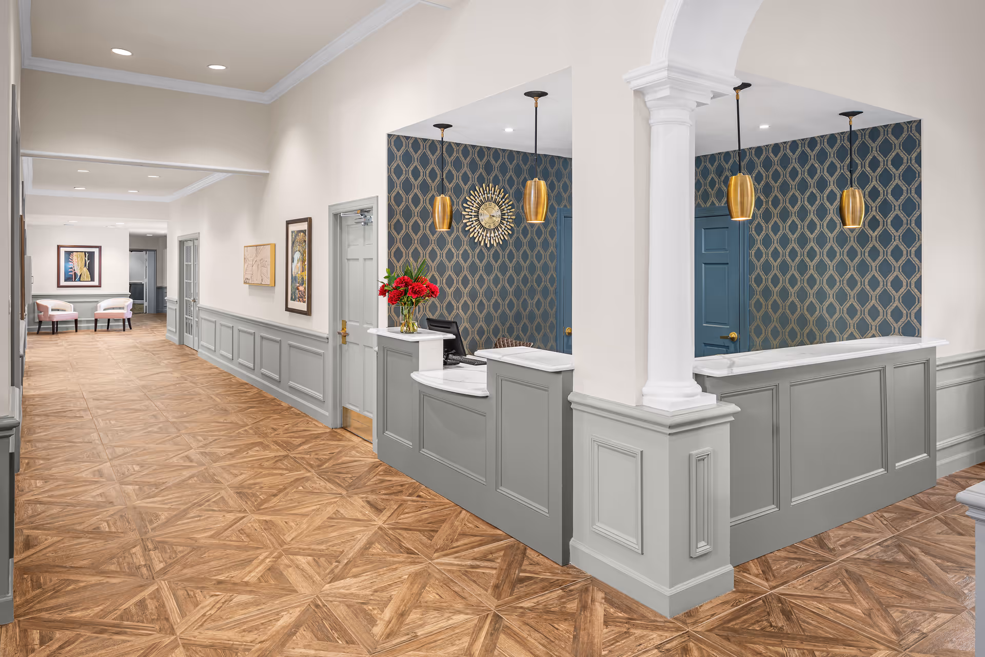 Interior view of a senior living facility reception area with a gray paneled front desk, two gold pendant lights, and a decorative blue and gold patterned wall behind the desk. The hallway features wood-patterned flooring, light gray wainscoting, and framed artwork on the walls. Two pink chairs and a framed picture are visible at the end of the hallway.