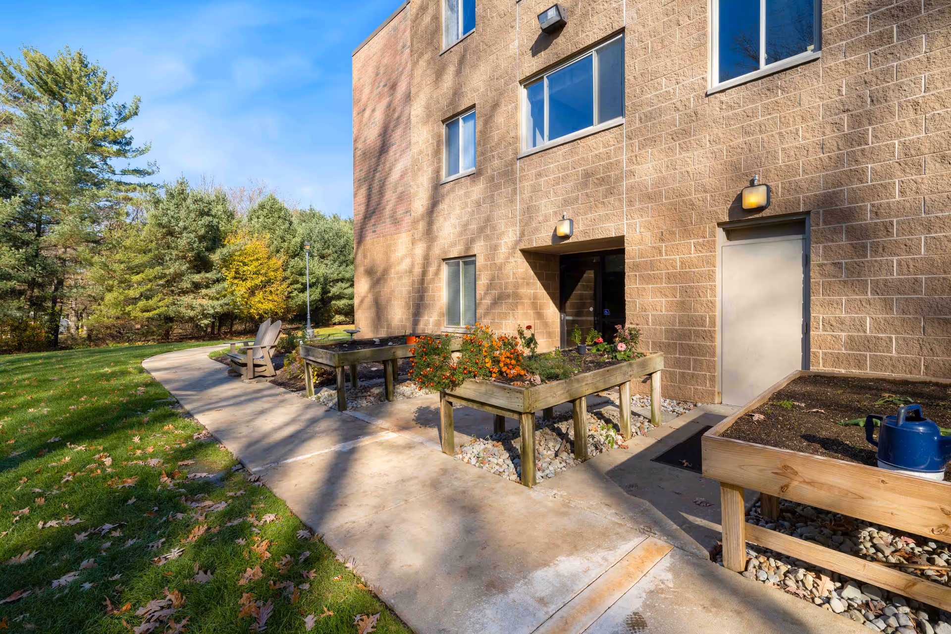 Outdoor garden area next to a brick building with raised wooden planter boxes containing flowers and soil, a blue watering can, a concrete pathway, and two wooden chairs facing a wooded area with green trees under a blue sky.