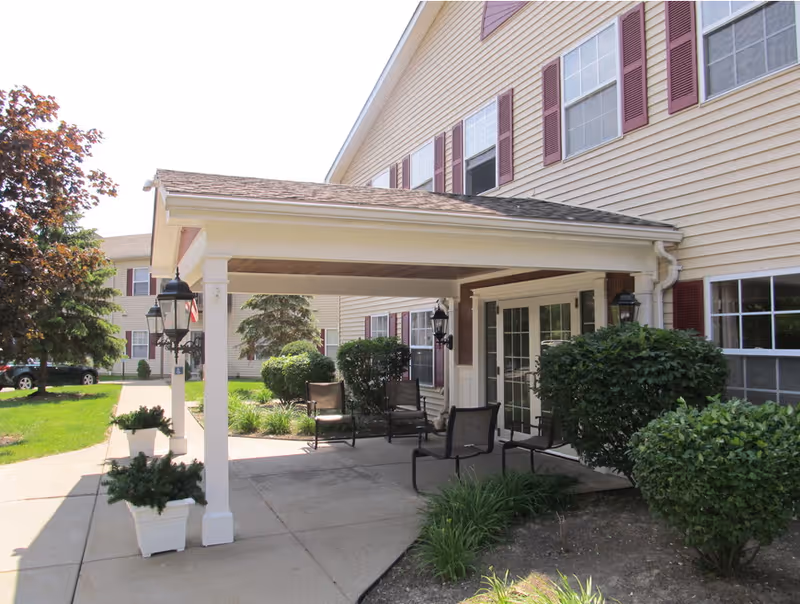 Covered entrance with chairs, potted plants, and shrubs at the front of a beige senior apartment building with maroon shutters.