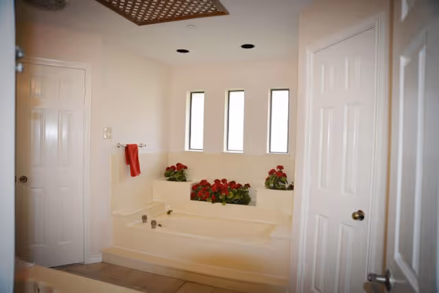 A bright bathroom featuring a large built-in bathtub with red flowers placed on the ledge behind it. There are three narrow vertical windows above the tub allowing natural light in. A red towel hangs on a towel rack on the left wall. The room has white walls and two white doors, one on each side of the tub.