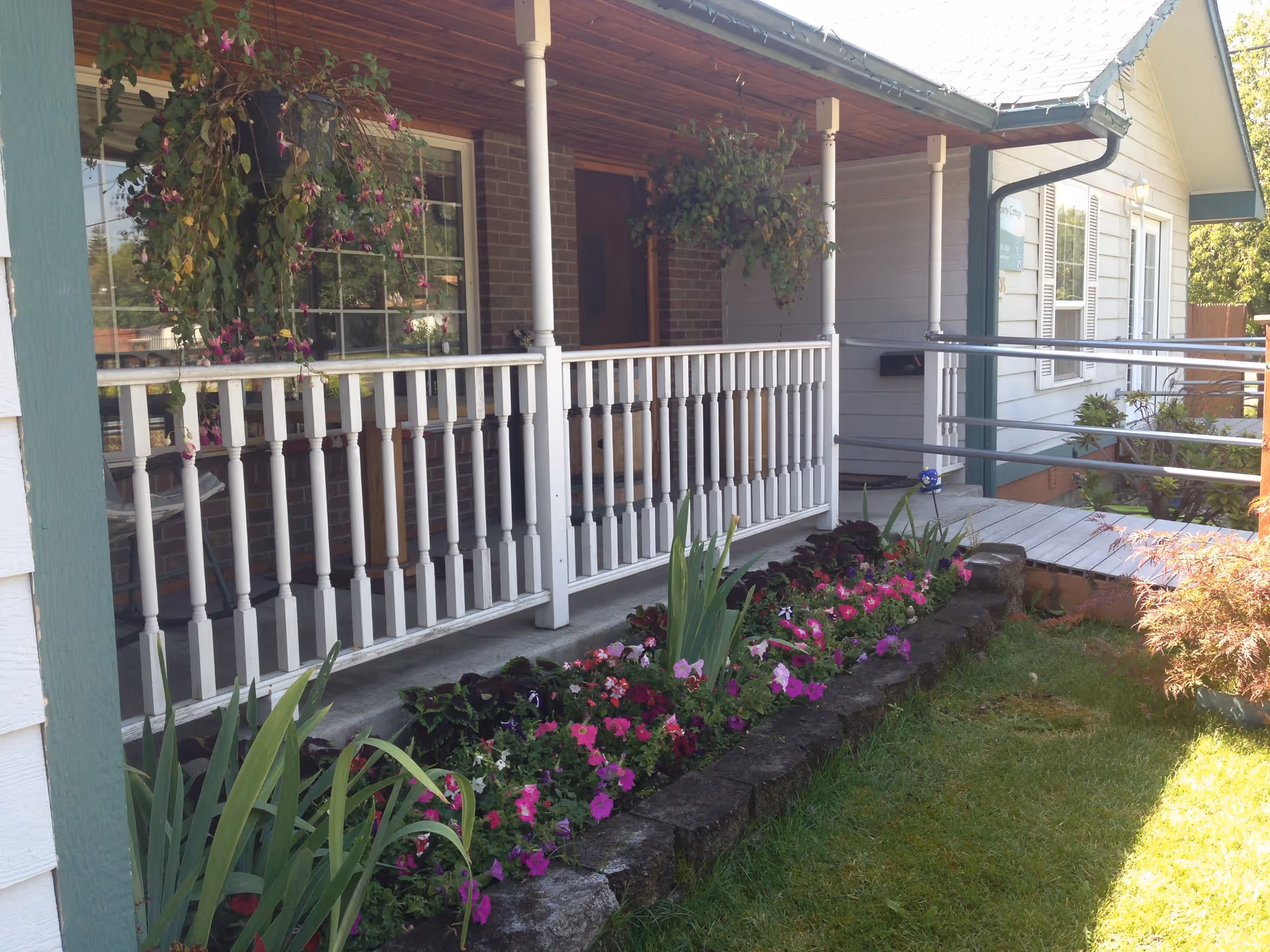 Front porch of a single-story building with white railings and hanging flower baskets. There is a flower bed with colorful flowers along the edge of the porch and a wooden ramp leading to the entrance. The building has white siding with green trim and a brick section near the door.