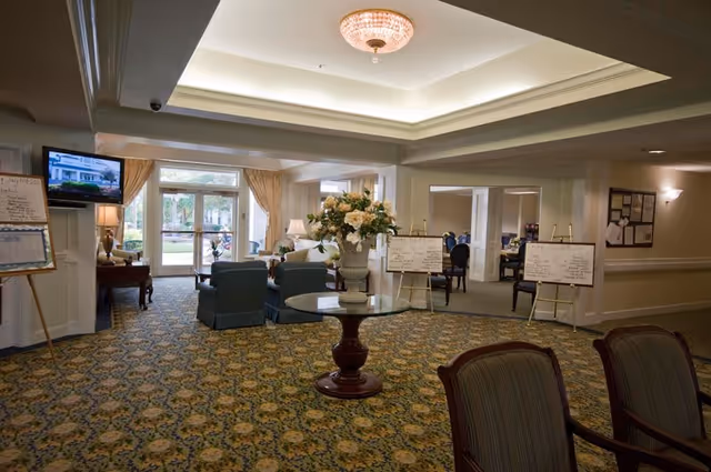 A spacious senior living facility common area with patterned carpet, a round table with a large floral arrangement in the center, several upholstered chairs, and large windows letting in natural light. There are whiteboards on easels with writing, a television mounted on the wall, and a view into adjacent rooms with more seating.