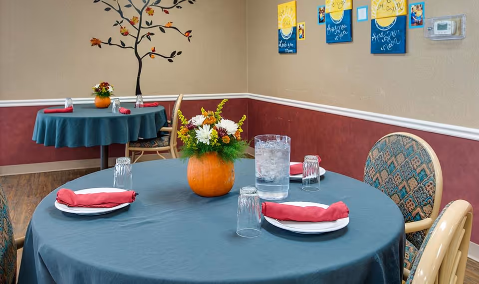 Small dining room with round tables set with plates, red napkins, glassware, and pumpkin flower centerpieces.