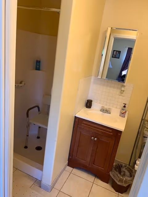 Small bathroom with a walk-in shower equipped with a white shower chair and a grab bar. Next to the shower is a wooden vanity with a white sink, a soap dispenser, and a small decorative item. Above the sink is a rectangular mirror reflecting part of an adjacent room. The floor is tiled, and there is a trash bin beside the vanity.