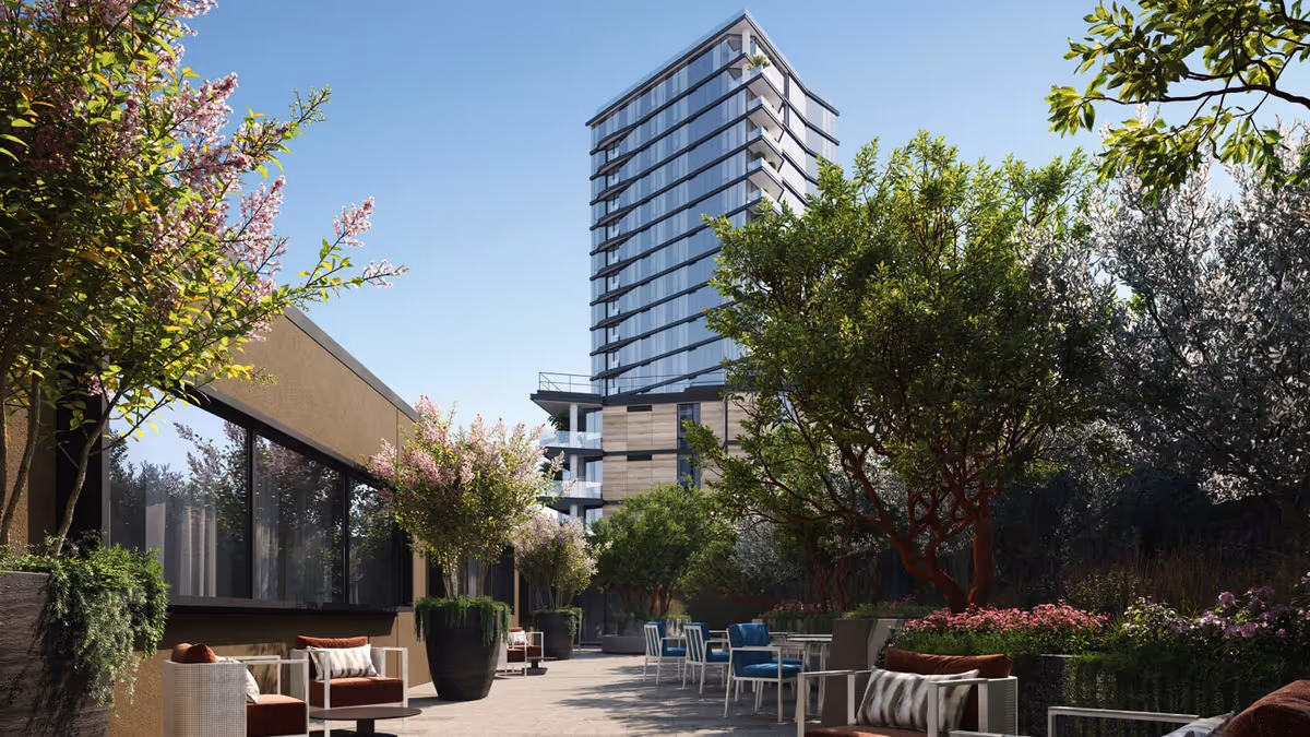 Outdoor patio area with modern seating arrangements including chairs and sofas surrounded by lush greenery and flowering plants, with a tall glass building in the background under a clear blue sky.