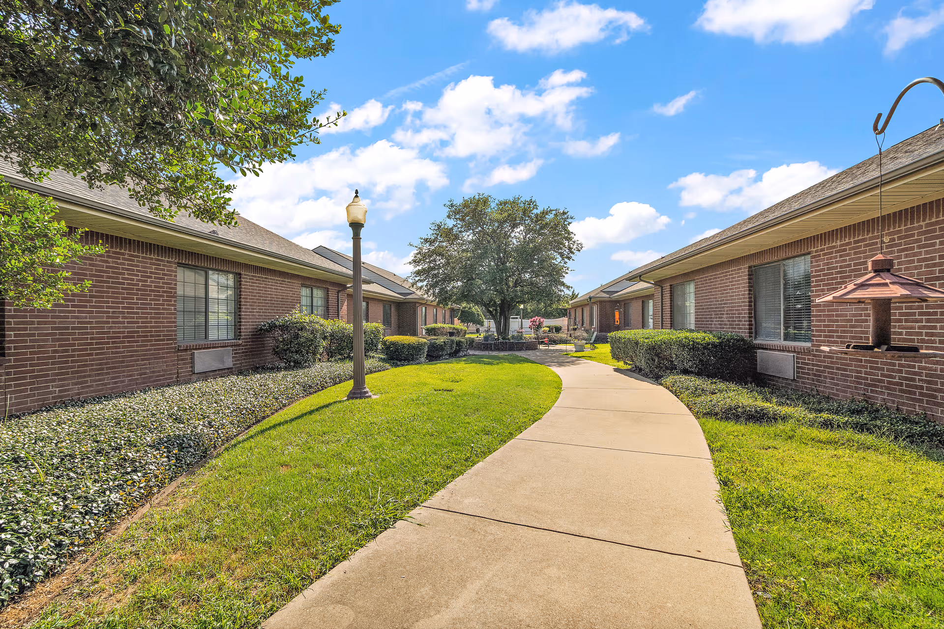 A paved walkway curves through a well-maintained grassy area between two single-story brick buildings with windows. There are bushes and small trees lining the path, a lamppost on the left side, and a bird feeder hanging on the right. The sky is blue with scattered clouds.
