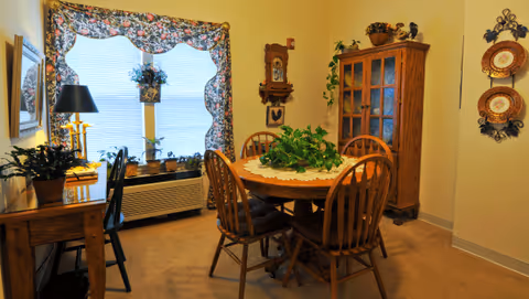 A cozy dining room with a round wooden table surrounded by four wooden chairs. The table has a white lace doily and a green leafy plant centerpiece. To the left, there is a wooden desk with a black chair, a lamp, and a potted plant. A large window with floral curtains lets in natural light. On the right side, there is a wooden cabinet with glass doors and decorative items on top. Two decorative plates hang on the wall beside the cabinet.