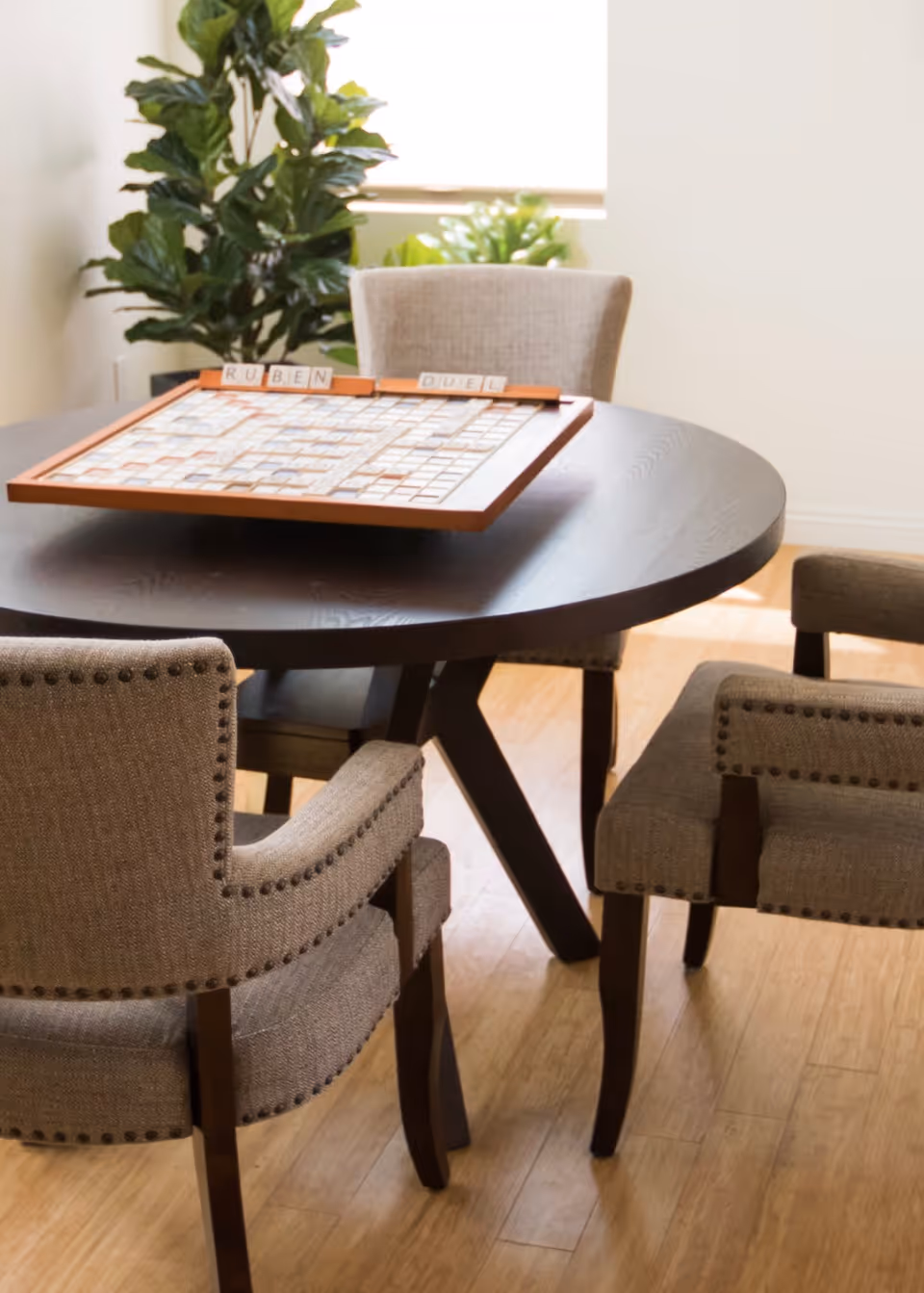 A round dark wooden table with a Scrabble board game set up on it, surrounded by four upholstered chairs with nailhead trim. There are two green plants near a window in the background, and the floor is light wood.