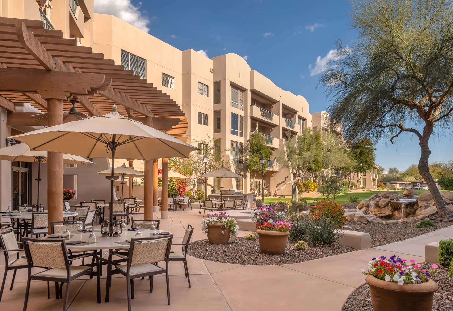 Outdoor patio area at Vi at Grayhawk with tables and chairs under large beige umbrellas, surrounded by potted flowers and desert landscaping. The building is a multi-story beige structure with balconies, and there is a small rock waterfall feature with trees and shrubs nearby under a clear blue sky.