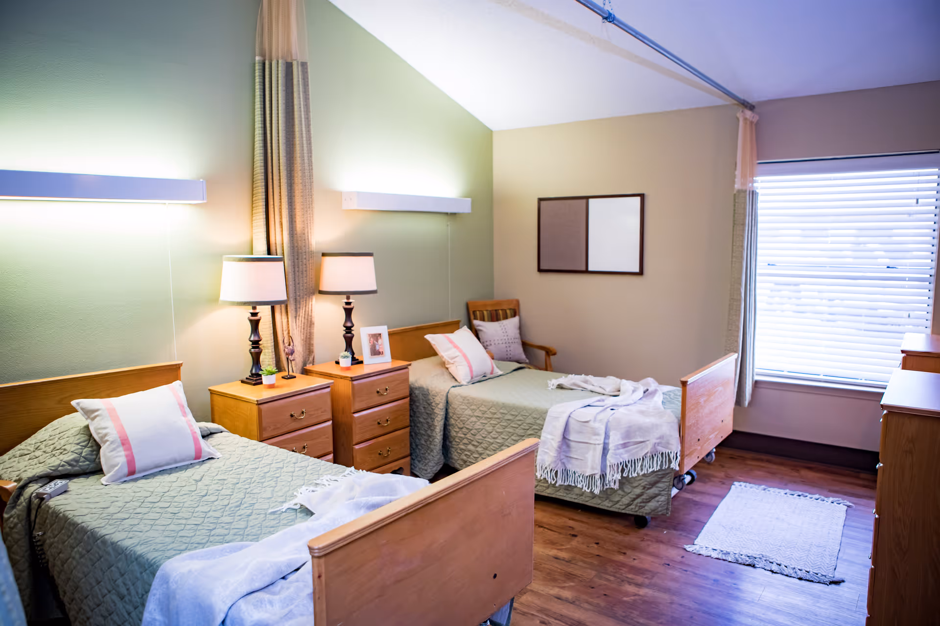 A bright and clean nursing facility bedroom with two single beds, each with green quilted bedspreads and pillows. Between the beds are two wooden nightstands with table lamps, a small plant, and a framed photo. The room has wooden flooring, a window with blinds, and a small rug near the window. The walls are painted in light green and beige tones, and there is a curtain divider between the beds.
