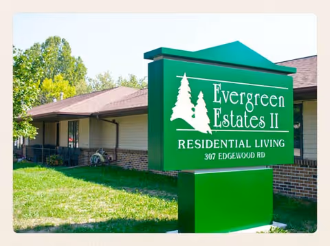 Green sign for Evergreen Estates II Residential Living at 307 Edgewood Rd in front of a single-story building with a brick and siding exterior, surrounded by grass and trees.