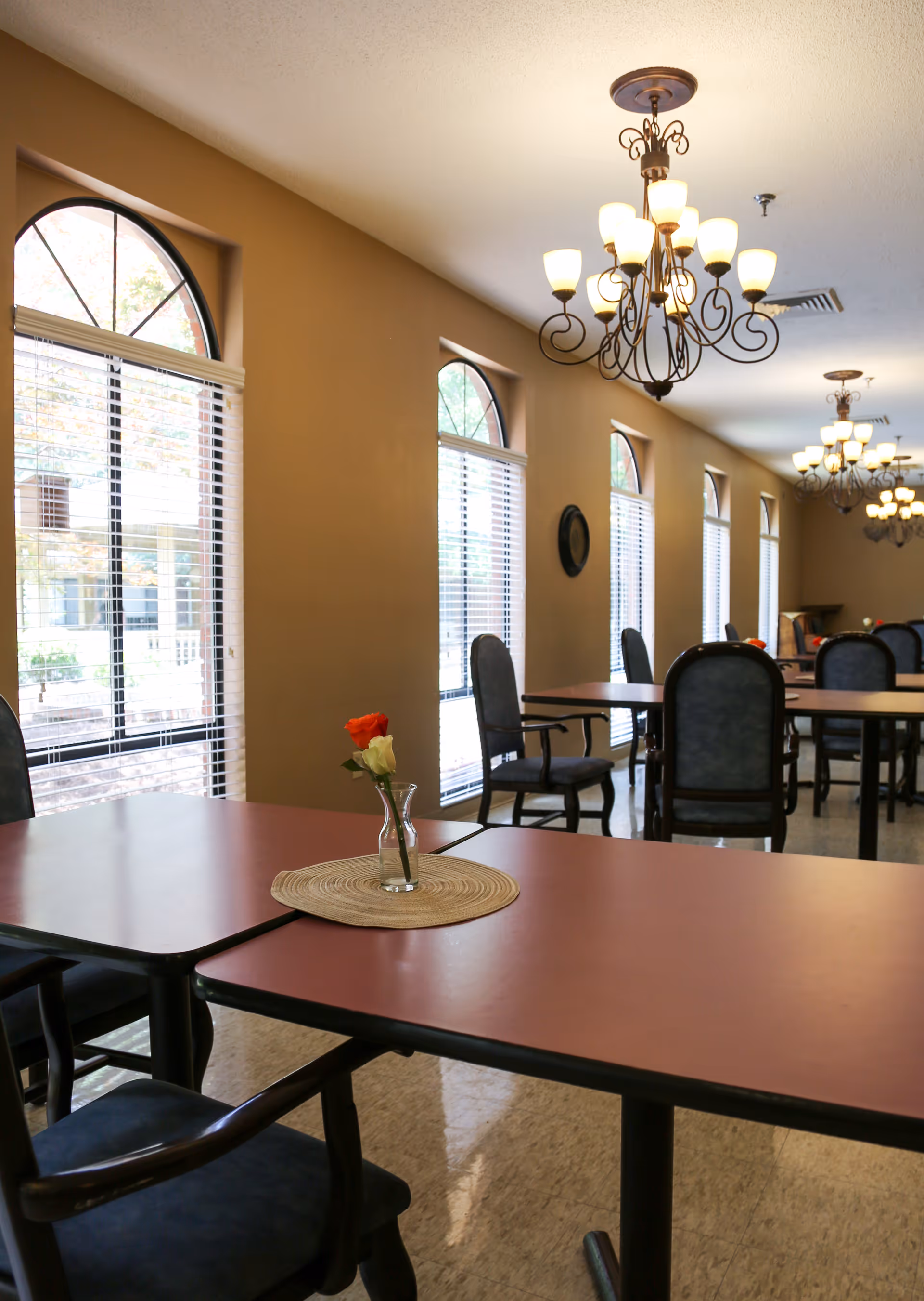 Dining room with tables and chairs, arched windows, chandeliers, and a small vase with a single rose on the foreground table.