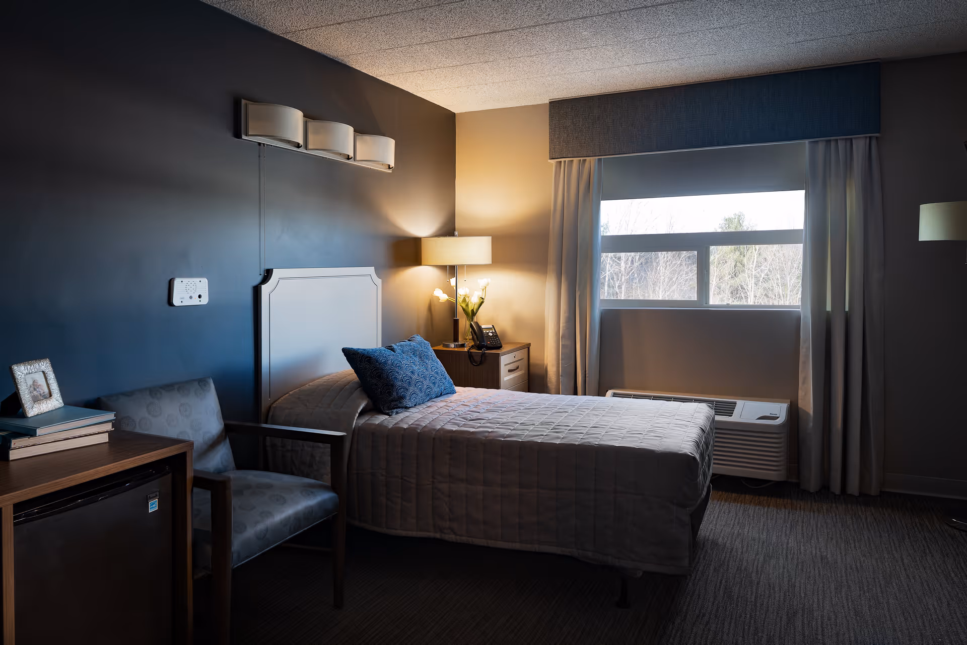 A cozy bedroom with a single bed covered in a beige quilt and a blue decorative pillow. Next to the bed is a wooden nightstand with a lamp, a telephone, and a small vase with flowers. A window with curtains lets in natural light, and there is a chair and a small refrigerator beside a wooden table with books and a framed photo. The walls are painted in dark and light tones.