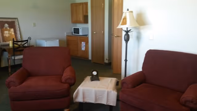 A living area with two red upholstered armchairs facing a small table covered with a white cloth and a small clock on top. In the background, there is a wooden cabinet with a microwave and a mini fridge, a floor lamp, and a small table with a chair and framed artwork on the wall.