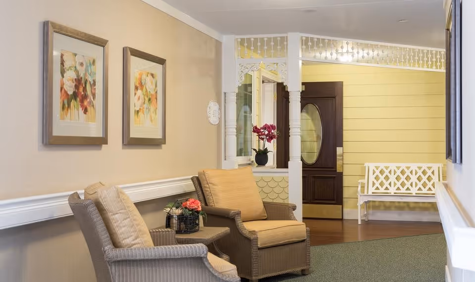 A cozy sitting area in a senior living facility hallway with two cushioned wicker chairs and a small table with a flower arrangement. The walls are decorated with two framed floral paintings. In the background, there is a dark wooden door with an oval window and a white bench against a yellow paneled wall.