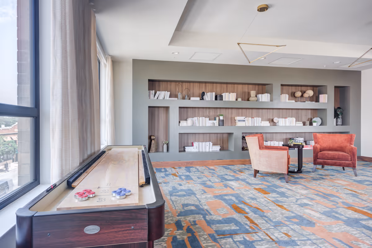 Bright communal lounge with a shuffleboard table by a large window, built-in bookshelves, and two upholstered chairs around a small table.