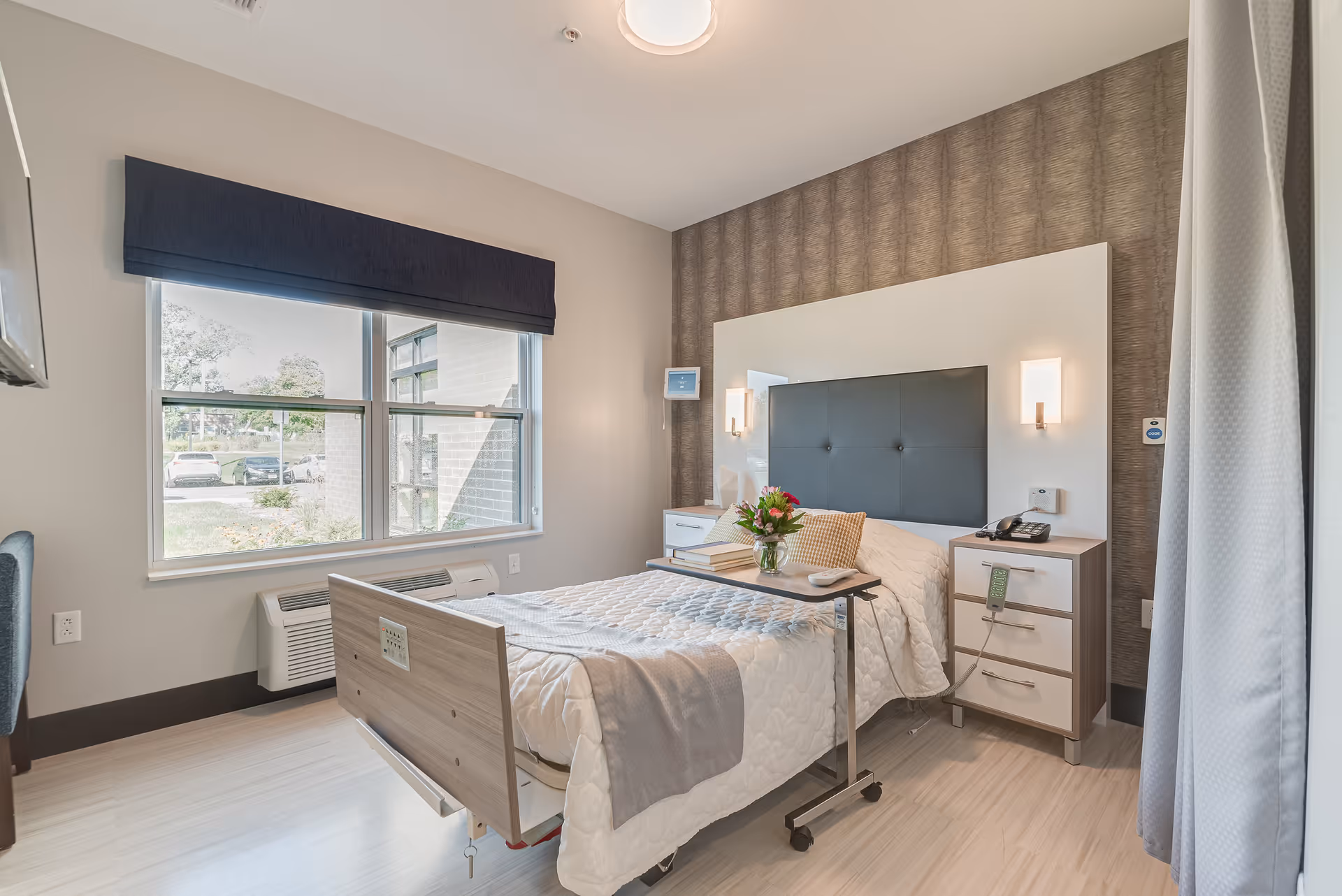 A bright and clean medical facility bedroom with a hospital bed covered in a white quilted blanket and a gray throw. There is a small overbed table with a vase of flowers and books. The room has a large window with a dark blue valance, a bedside table with a phone and medical devices, and a textured accent wall behind the bed.